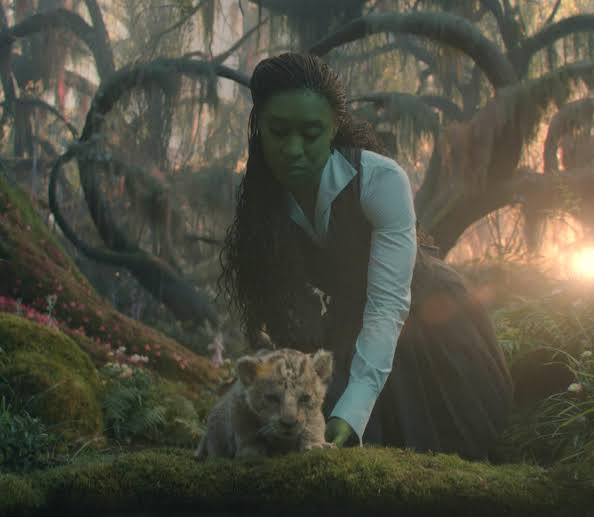 Elphaba kneeling beside a lion cub in a forest.